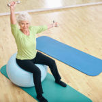 35084417 - senior woman sitting on a pilates ball and lifting dumbbells looking at camera. old caucasian woman exercising with weights at gym.